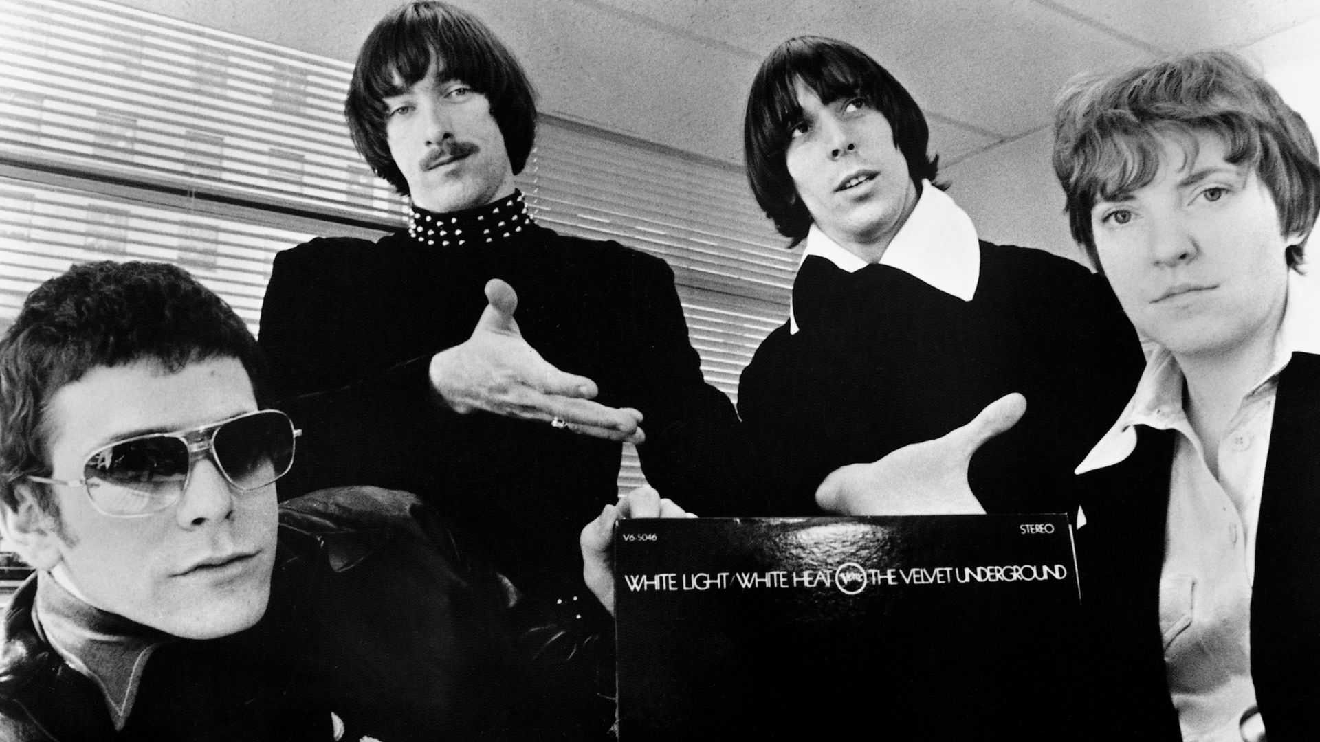 A publicity photo of the American rock band The Velvet Underground circa 1968, promoting their second album White Light/White Heat. The band members are positioned around a copy of the album, with the text of the title visible against a black background. From left to right: Lou Reed, Sterling Morrison, John Cale, and Maureen Tucker.
