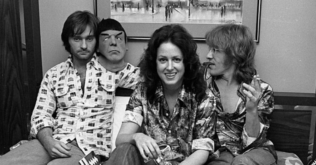 Marty Balin, Grace Slick, and Paul Kantner of Jefferson Starship seated on hotel room bed with a Mr. Spock mask in Los Angeles, California